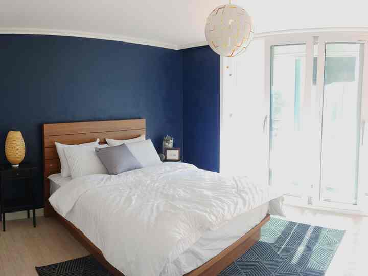 ネイビーのアクセントウォールと木目調のベッドが配置された、明るい白い天井と大きな窓が印象的な新婚の寝室