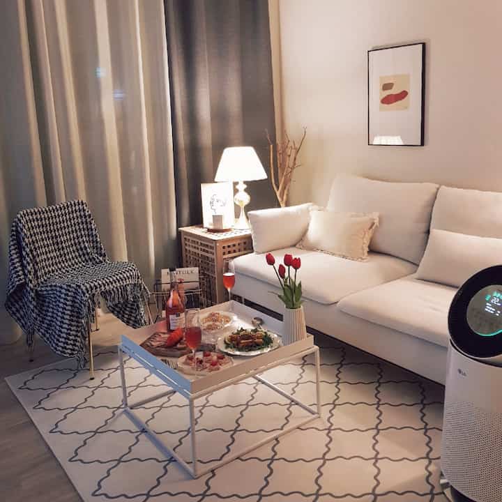 Bright white and beige living room featuring white sofa, coffee table, blackout curtains and warm lighting creating cozy atmosphere