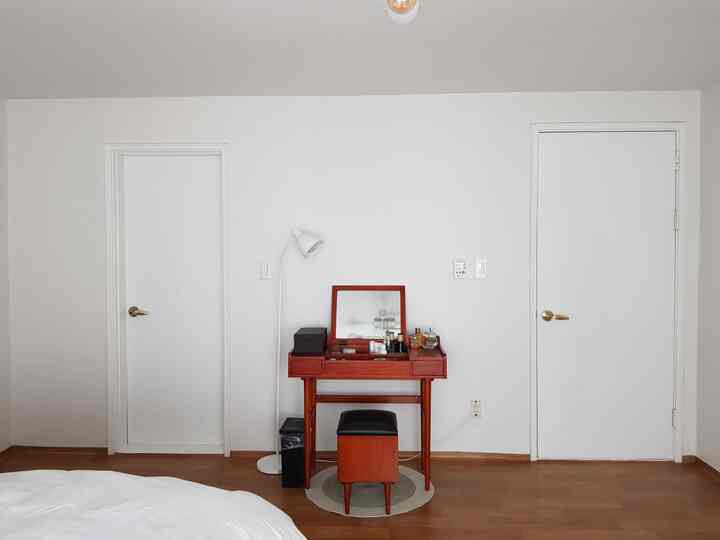 White and brown toned bedroom wall featuring a wooden vanity and stool centered, with restroom door and floor lamp positioned left, creating a simple space