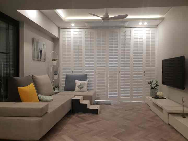 A white and beige toned living room featuring large louver shutters, herringbone floor, sectional sofa, and ceiling fan for a cozy atmosphere