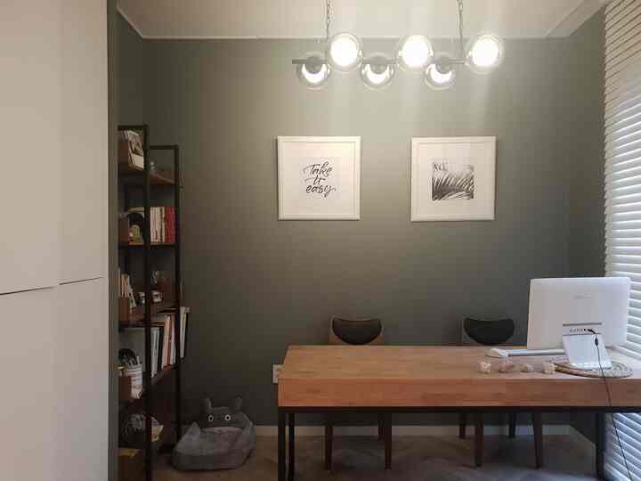 オリーブグリーンの壁とナチュラルウッドのデスクが配置された書斎空間、ブラックのペンダントライトと棚が調和した落ち着いたインテリア