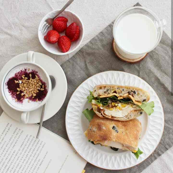 A natural-toned table setting featuring an olive ciabatta sandwich, strawberries, yogurt, and milk in a modern, clean breakfast scene