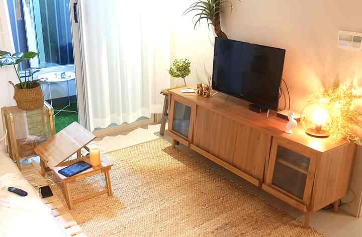 ナチュラルなウッドトーンとベージュのリビングルーム、ラタンカーペットと木製テレビ台、自然光と植物が調和した落ち着いた空間