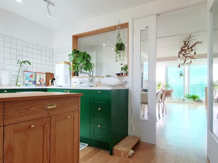 Bright white kitchen and dining area with wood tone floors, green cabinets and plants adding a natural atmosphere