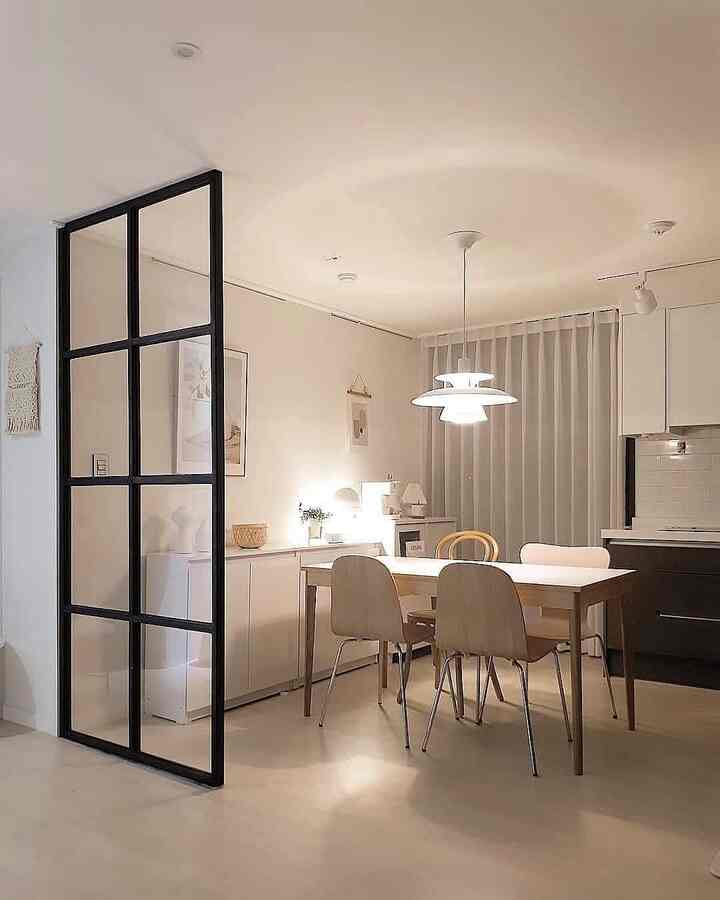 Simple dining room with white and wood tones, featuring a pendant light and black-framed room divider creating a modern, open atmosphere