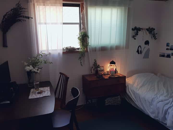 Studio bedroom with wood tone furniture and white bedding, featuring curtains and plants by windows in a cozy setting