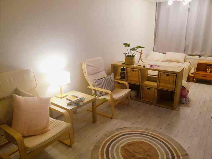 White and light brown toned long and narrow studio featuring armchairs and a bed separated by a wooden divider with a natural, cozy atmosphere