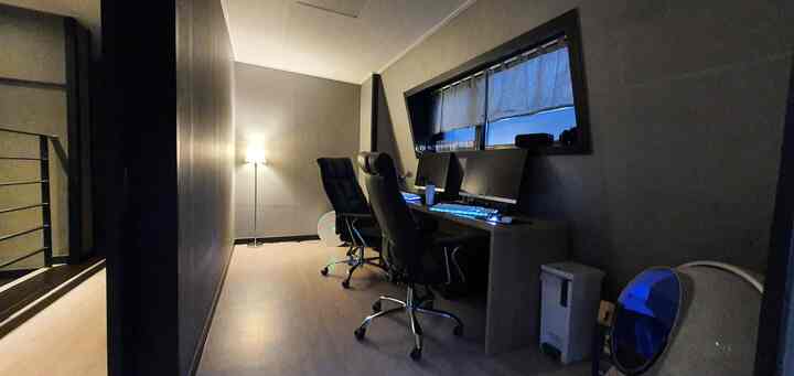 Gray-toned walls and black gaming chairs in a narrow, compact study-style game room featuring a long desk and floor lamp creating a cozy atmosphere
