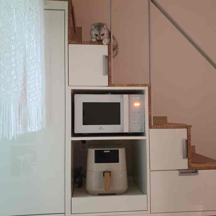 White and beige toned duplex officetel kitchen storage space featuring a microwave, air fryer, and a cat atop the staircase with a lace curtain