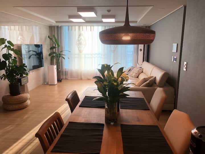 Beige natural-tone living room featuring wooden dining table and chairs with large tropical plants creating a cozy atmosphere