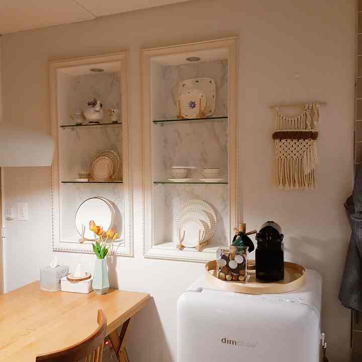 White and wood tone kitchen space featuring a wooden dining table and chair, ceramic dishes on shelves, and a coffee machine creating a natural atmosphere
