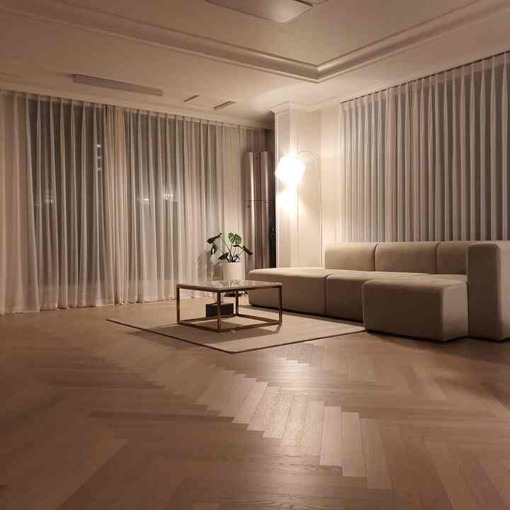 White and wood tone living room for two featuring herringbone flooring, simple sofa, curtains, and floor lamp with cozy ambiance