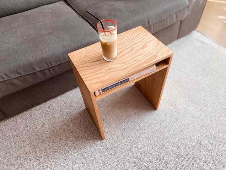 Natural wood-toned nightstand placed on beige rug beside a gray sofa, featuring a coffee glass on top in a cozy living room setting