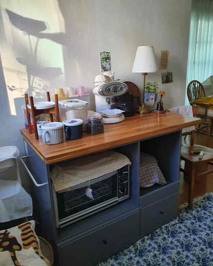 明るい木目調の天板とグレーの脚部を持つキッチンアイランドが中心のナチュラルなキッチンスペース、ホームカフェのコーヒーマシンとフロアランプが配置された居心地良い雰囲気