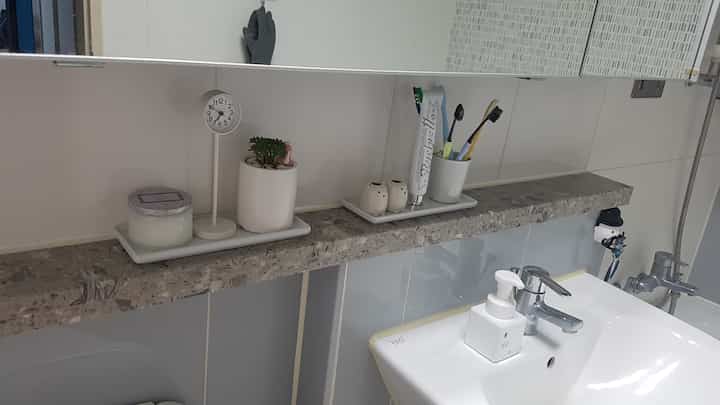 グレーの大理石の棚に配置された白いトレイ、キャンドル、小さな多肉植物、歯ブラシ立てがあるバスルームの洗面台エリア