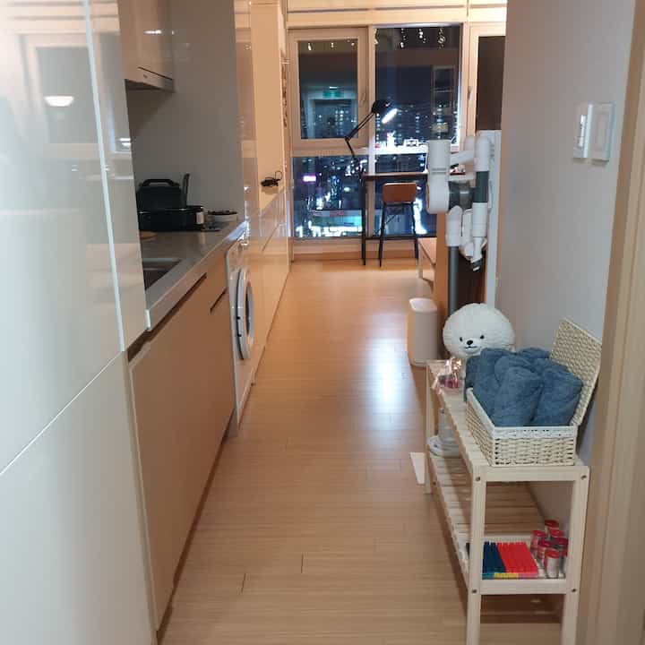 明るい木目調の床と白いキャビネットが特徴の細長いキッチン空間、窓際にホームオフィス家具とタオル収納が配置された整ったインテリア