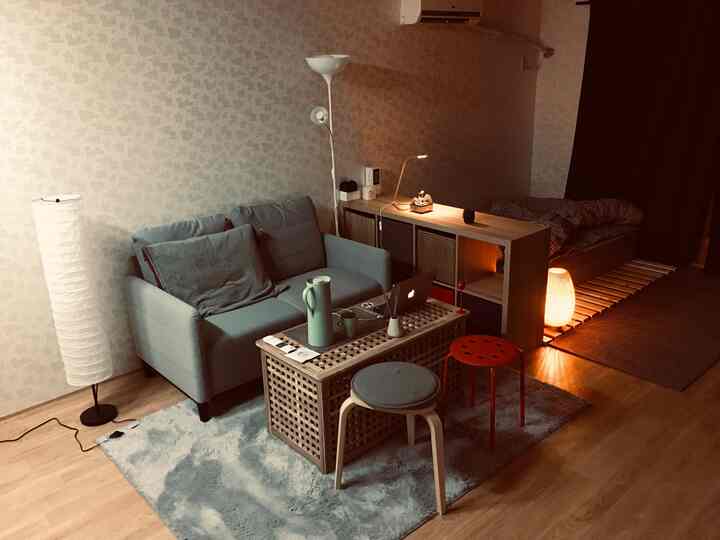 A long, narrow studio apartment with white walls and natural wood flooring, featuring a blue sofa, stools, and a KALLAX shelf dividing bedroom and living area, cozy atmosphere