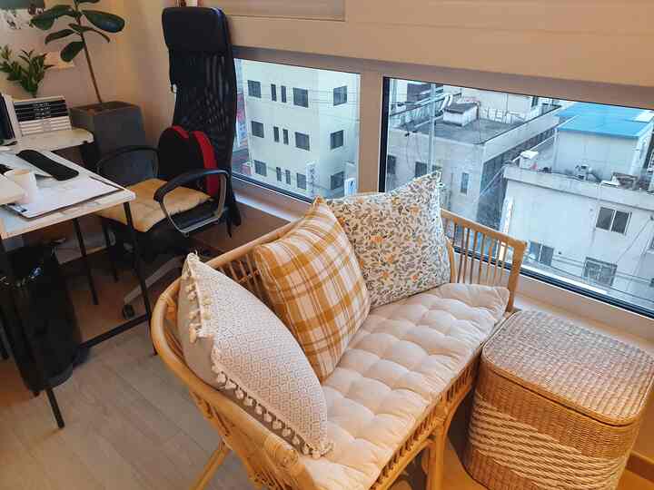 Natural tone compact home office space featuring rattan 2-seater sofa with cushions, desk, and chair beside a large window, creating a cozy atmosphere