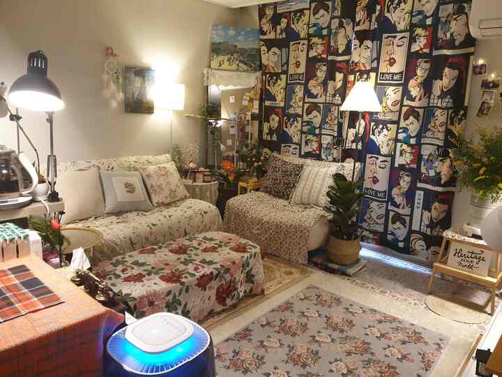 暖かい照明とフロアランプが配置された小さなリビング、花柄のソファとポップアートのカーテンが映える雰囲気のあるインテリア