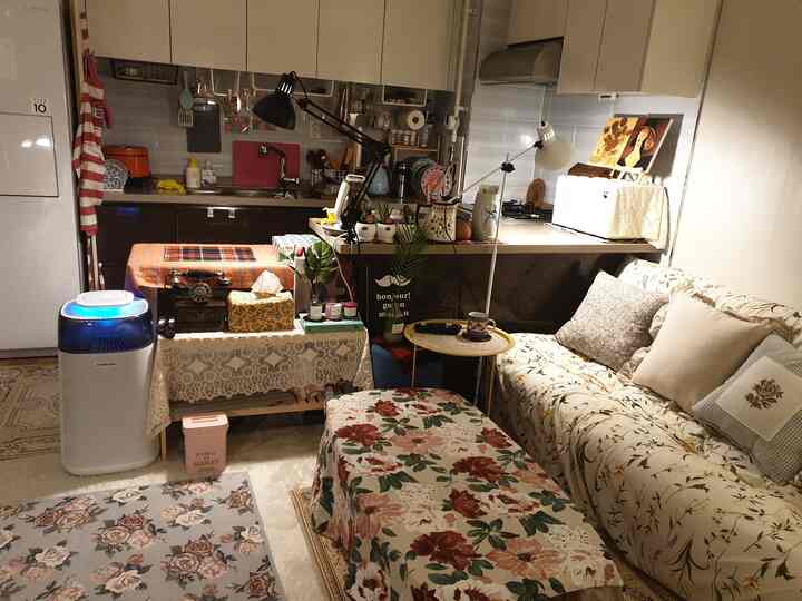 温かみのあるホワイトとブラウンの色調のリビングとキッチン、フロアランプと植物、パターンソファが配置された雰囲気のあるインテリア