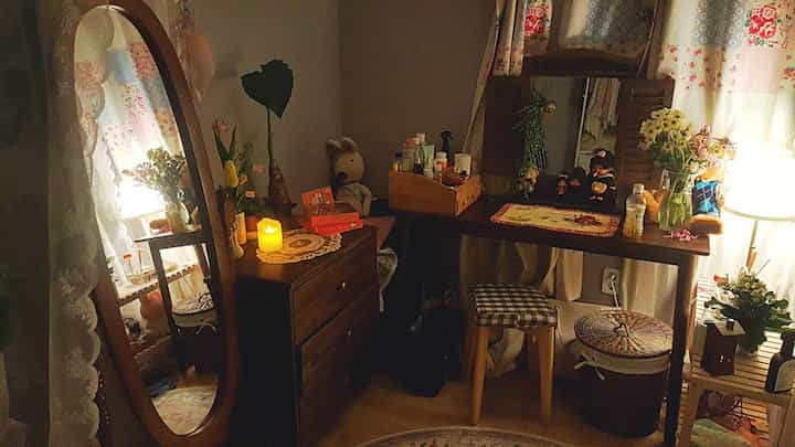 温かみのある木目調の寝室、ナイトスタンド、鏡、植物、ヴィンテージ風の装飾品が配され、レースの花柄カーテンが調和する落ち着いた空間