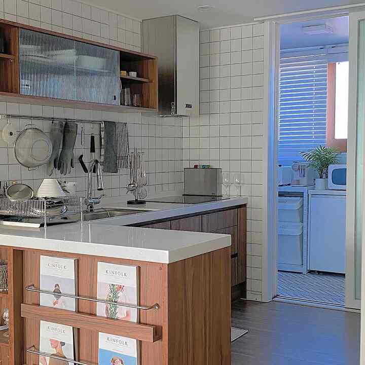 白いタイル壁と木目調のアイランドキッチンが配置された、雑誌ラックとキッチン用品があるモダンな印象の空間