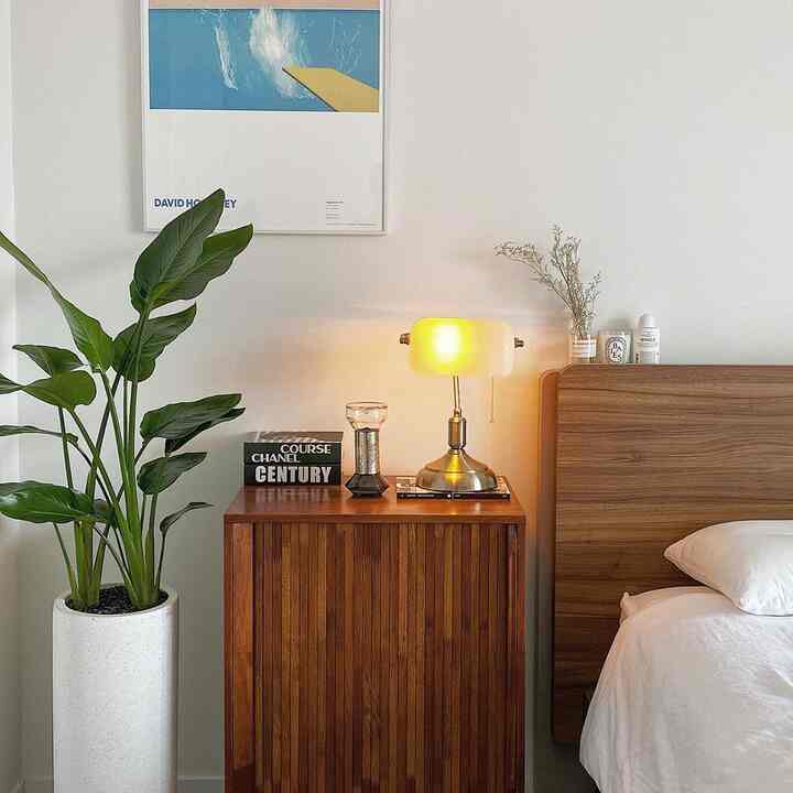 白と茶色のトーンの寝室、背の高い植物と木製ナイトテーブルに温かい光のテーブルランプとデイヴィッド・ホックニーの額が配置された自然な雰囲気のあるインテリア