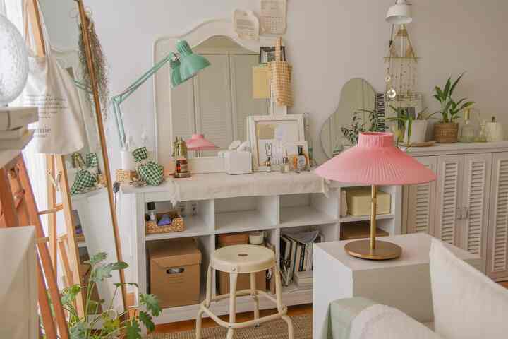 白を基調としたシンプルでナチュラルな印象の小さな部屋、化粧台と収納家具、ピンクのテーブルランプが配置されている