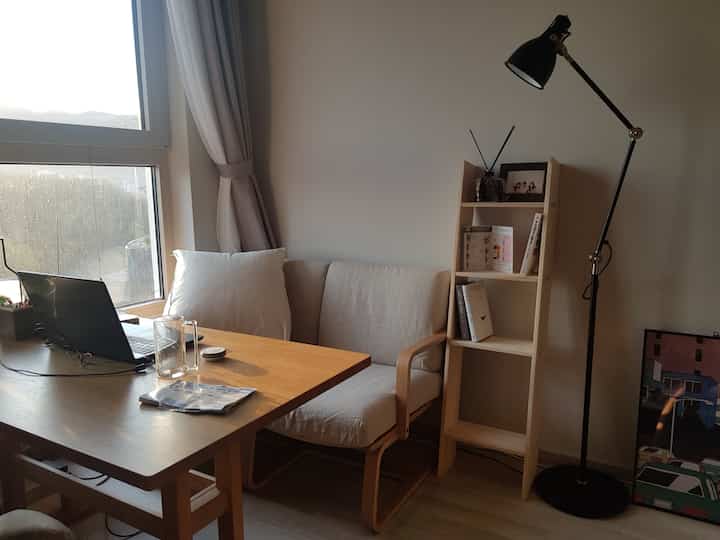 ナチュラルな木目調とベージュのソファがあるコンパクトなワンルームのリビング、フロアランプとラックが配置された落ち着いた空間