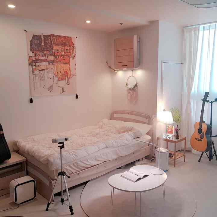 Ivory and white-toned studio bedroom featuring a wooden bed frame, folding table with an open book, macrame and poster adding warm, cozy atmosphere