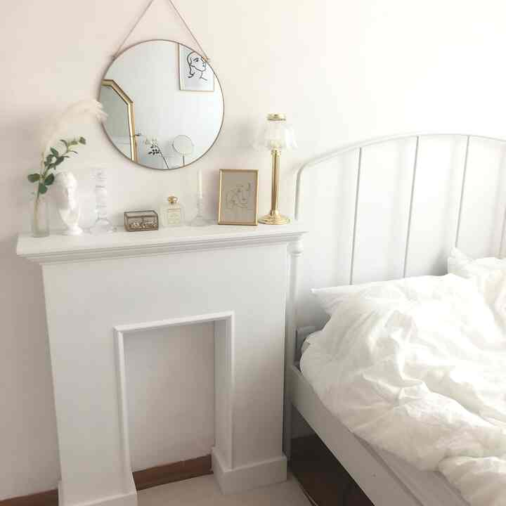 White and gold toned bedroom featuring a decorative fireplace with a gold table lamp in the center and a metal bed on the right, showcasing a clean interior design
