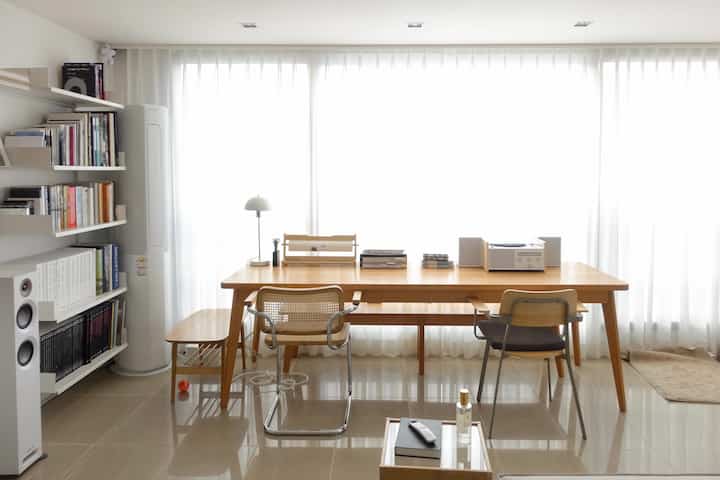 Natural-toned study space featuring a long wooden table, armchairs, and bookshelf arranged in a clean, bright living room corner