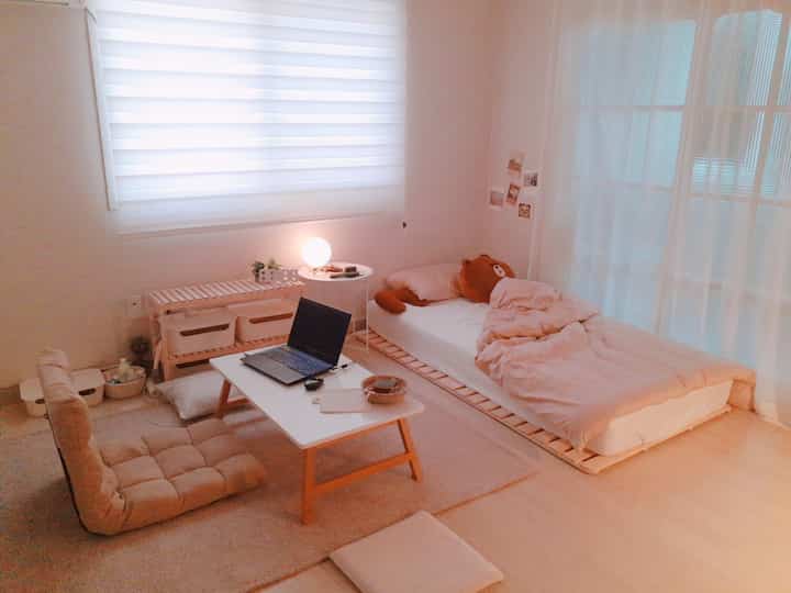 Warm beige natural tone studio bedroom featuring floor chair, low table, bed, and sheer curtains in a cozy setting