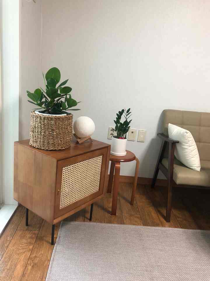 ナチュラルな木目調のリビングの一角、ラタンのナイトテーブルと木製スツールに植物とムーンランプが配置されたシンプルな空間