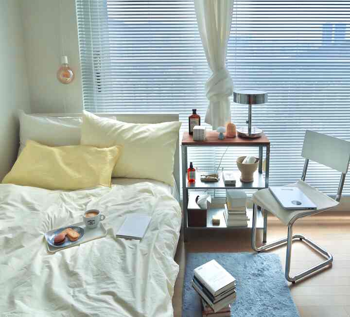 A white and natural toned bedroom featuring tidy bedding, a nightstand, and blinds in a simple, clean interior