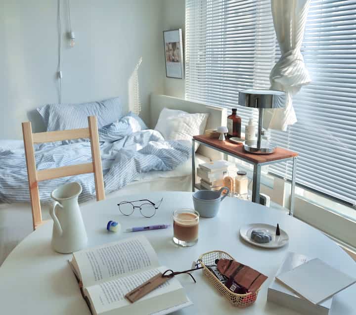 Bright bedroom in white and natural wood tones featuring blinds by the window and table lamp creating a cozy reading nook