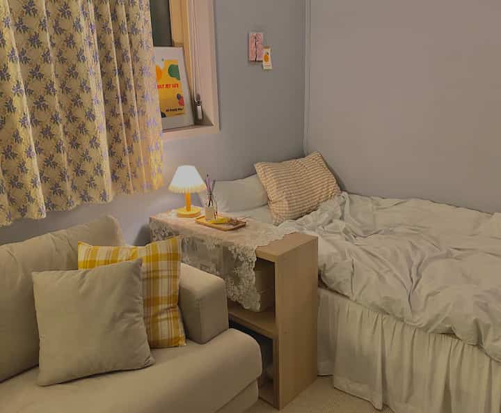 Beige and white toned studio bedroom featuring sofa, bed, floral curtains, and lace table runner in a cozy atmosphere
