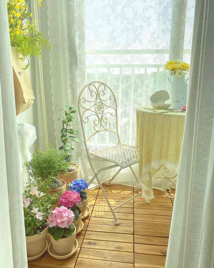 White and wood tone veranda featuring French country style iron chair, lace curtains, and various potted plants creating a cozy atmosphere