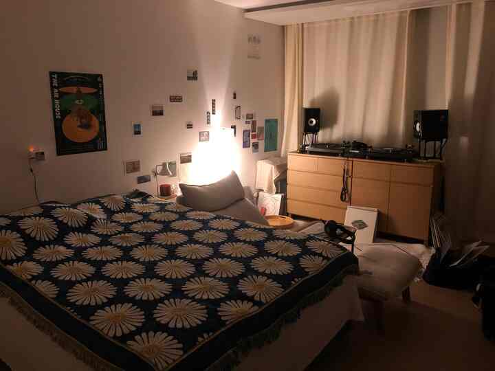 ナチュラルな木材家具と白いカーテンがあるミニマルな寝室、フラワー柄のベッドカバーとオーディオシステムが印象的な空間