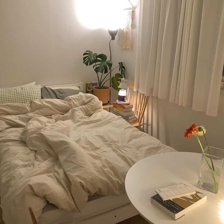 Soft white-toned small studio bedroom featuring wooden nightstand, plant, and floor lamp creating a cozy atmosphere
