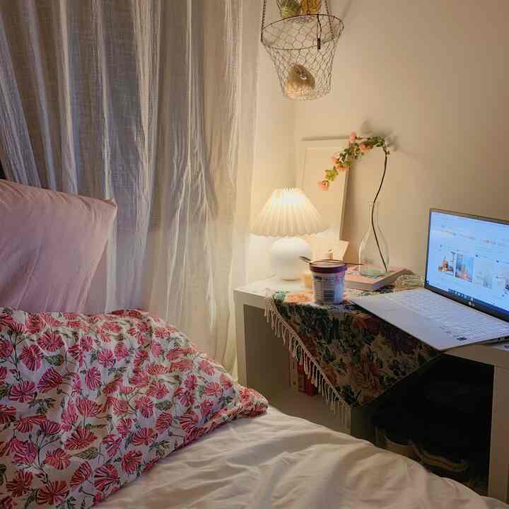柔らかいホワイトとピンクの色調の小さな寝室、花柄の寝具とノートパソコンが置かれたデスクが配置された雰囲気のあるヴィンテージ風インテリア