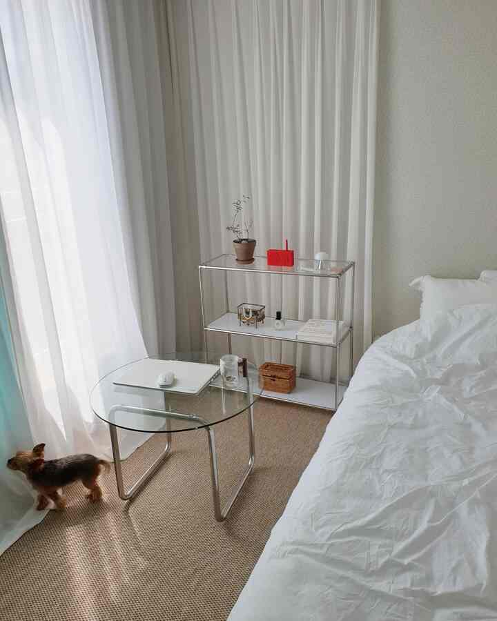 ナチュラルなトーンの小さな寝室、白い寝具とカーテン、ガラステーブルとラタンの棚が配置された雰囲気のあるインテリア、小型犬が窓際にいる