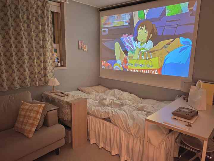 ナチュラルな色合いのワンルーム寝室、ソファとベッドが並び、プロジェクタースクリーンで映画が映し出される雰囲気のあるインテリア