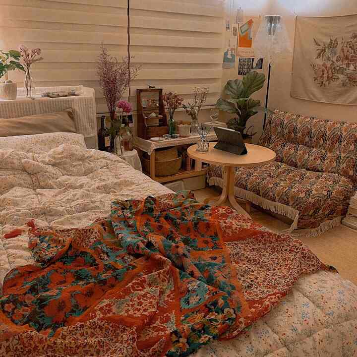 A small beige and natural toned bedroom featuring a vintage patterned sofa, round table, plants, and cozy bedding creating a comfortable atmosphere