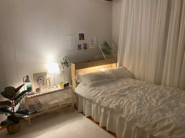 柔らかなホワイトトーンとウッドカラーが調和した印象的な寝室、枕と布団が置かれたベッドと間接照明のテーブルランプが配置された空間