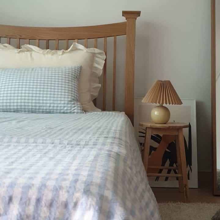 Natural wood tone and white bedroom featuring wooden bed, checked bedding, and a table lamp on a stool creating cozy atmosphere