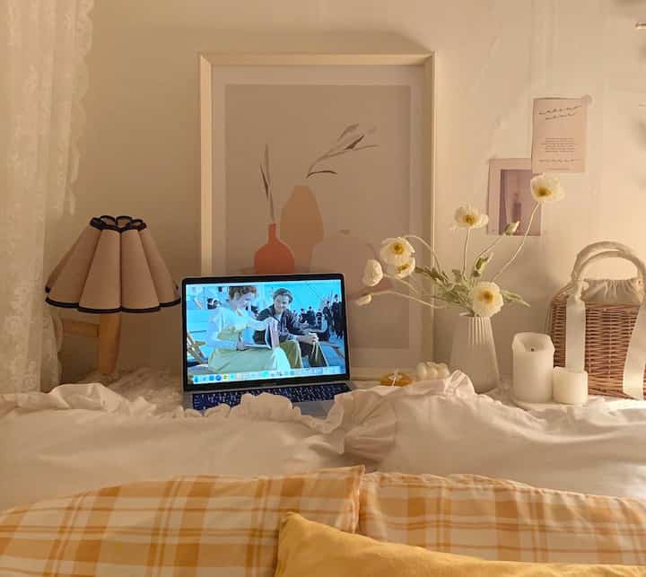 Warm beige and white toned bedroom featuring a bed with laptop, vase with flowers, and candles creating a cozy atmosphere