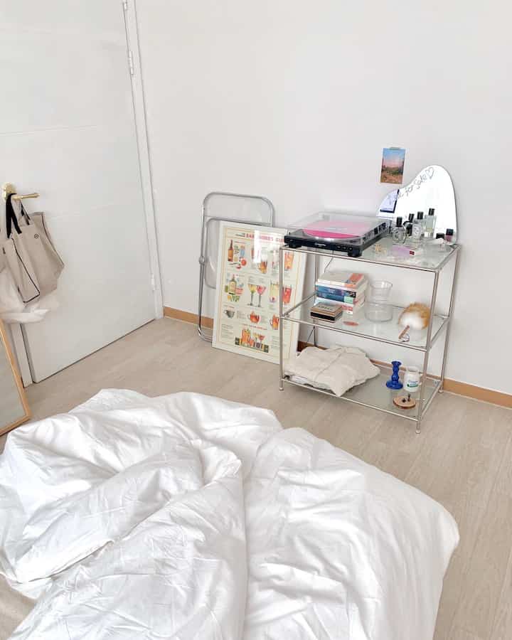 白い壁と床が際立つ寝室、透明なメタルのナイトテーブルにピンクのターンテーブルと様々な装飾品が配置された清潔感のある空間