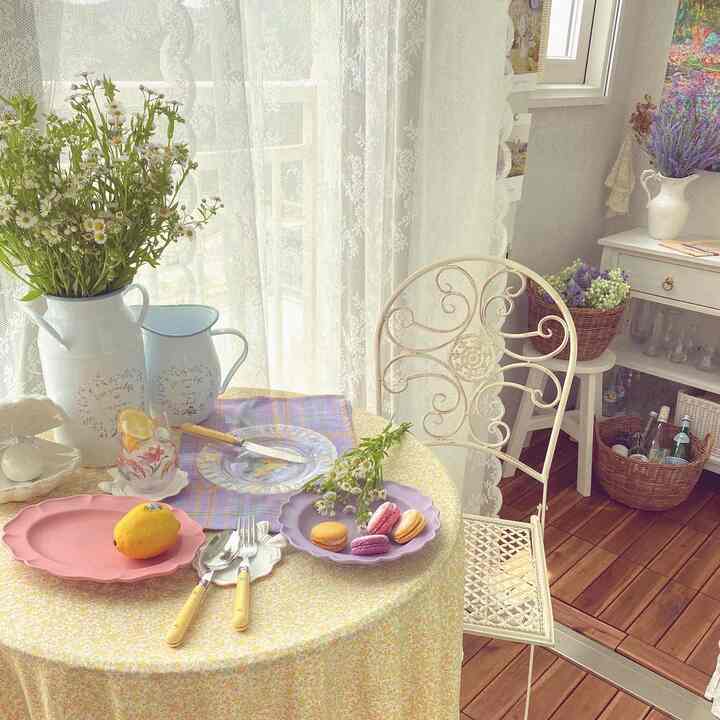 A balcony space featuring a yellow floral tablecloth on a round table, white wrought iron chair, lace curtains, pastel plates, and floral decorations in a cozy French Country style
