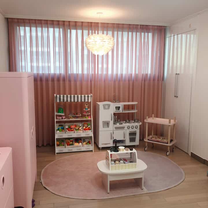 Pink-toned kids' room featuring pink curtains and rug, white play kitchen set, and toy storage for a cozy atmosphere